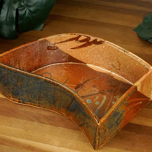 May include: A handmade, ceramic, corner-shaped dish with a rustic aesthetic. The dish features a blend of brown, orange, and blue hues, with subtle decorative markings. The design is ideal for displaying small items or as a decorative accent piece.