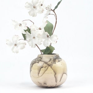 May include: A small, round, off-white ceramic vase with a map-like design, holding a stem of white flowers with green leaves. The vase has a unique, textured appearance, and the flowers add a touch of natural beauty.