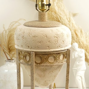 May include: A cream-colored table lamp with a textured, urn-shaped base and a gold-colored metal frame. The lamp has a light bulb and a beige lampshade. The lamp is on a white surface.