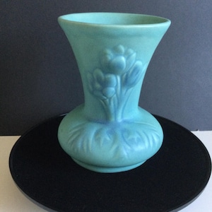 Peut inclure: Un vase turquoise en céramique avec un haut évasé et une base arrondie, orné d'un motif floral en relief. Le vase est posé sur un support noir circulaire. Environ 20 cm de hauteur.