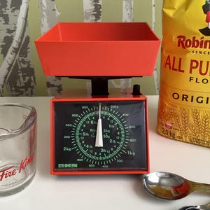 May include: A red kitchen scale with a black dial and a white pointer. The dial has markings for pounds, kilograms, and ounces. The scale is sitting on a white surface. The scale is labeled "EKS".
