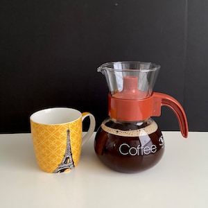 May include: A glass coffee pot with a red lid and handle, filled with coffee. The pot has the word "Coffee" printed on the side. A yellow mug with a black Eiffel Tower design is next to the pot.