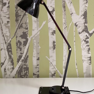 May include: A black desk lamp with a rectangular base and adjustable arm. The lamp has a conical shade and a dark burgundy arm. The lamp is on a white surface with a green and gray tree-themed wallpaper background.