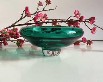 May include: A teal green glass bowl with a wide, shallow base and a small, round foot. The bowl is sitting on a white surface with a branch of pink blossoms.
