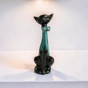 May include: A ceramic cat ornament with a long neck and a bow tie. The cat is mainly black with a teal green neck and accents. The figurine is sitting upright and has a glossy finish.