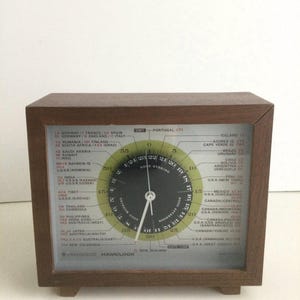 May include: A wooden framed clock with a world time zone dial. The dial has a clock face with hands and a ring of time zones around the outside. The clock face reads "Good Evening". The time zones are listed around the outside of the clock face. The clock is made by Kenwood and is called a "Hamclock".