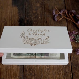 Könnte beinhalten: Eine weiße Holzbox mit Klappdeckel, auf dem die Namen "Christopher & Niamh" und das Datum "04/07/2025" in goldfarbenen Buchstaben stehen, umgeben von einem floralen Kranz. Die Box ist geöffnet und zeigt Geld.