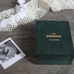 Op de afbeelding: Een donkergroene rechthoekige houten doos met goudkleurige gegraveerde tekst op het deksel met de tekst "Taufe Johanna 19. Juli 2026". Naast de doos ligt een vierkante zwart-witfoto van een baby, met een witte kanten rand in de buurt.