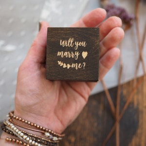 May include: A dark brown wooden ring box with the engraved phrase "Will you marry me?" in a gold color, along with a heart symbol and floral accents. The box is held in a hand, with bracelets visible.