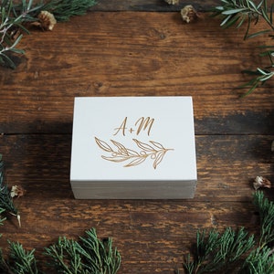 Op de afbeelding: Witte houten doos met een gouden gegraveerd ontwerp van een tak met bladeren en de initialen "A + M".