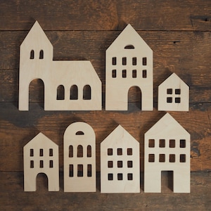 Puede incluir: Un conjunto de siete casas de madera recortadas en varios tamaños y estilos, dispuestas sobre una superficie de madera marrón. Las casas están hechas de madera de color claro y tienen formas geométricas simples.