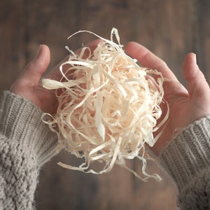 Può includere: Un paio di mani che tengono una grande quantità di trucioli di legno bianchi. I trucioli sono morbidi e soffici e vengono tenuti in posizione di coppa.