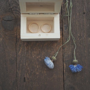 May include: Two gold wedding rings in a small wooden box with a hinged lid. The box is sitting on a dark brown wooden surface with two blue flowers.