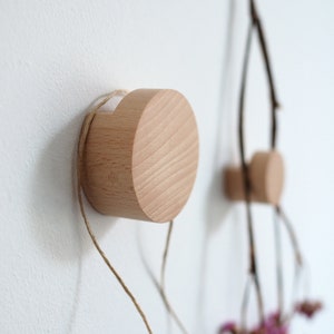 May include: Two wooden wall hooks with a natural finish are mounted on a white wall. The hooks are cylindrical with a flat front and a small hole for hanging. One hook has a natural twine attached. The other hook has a branch with small pink flowers.