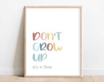 May include: A framed print with the words "DON'T GROW UP" in colorful, playful lettering. Below, the text "it's a trap" is written in a smaller font. The artwork is set against a white background, framed in a light wood.