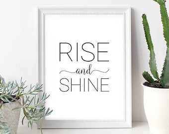 May include: A white framed print with the text "RISE and SHINE" in black lettering. The print is sitting on a white surface with two potted plants.