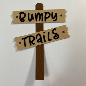 May include: A brown wooden sign with two beige signs attached to it. The top sign says "Bumpy" and the bottom sign says "Trails".