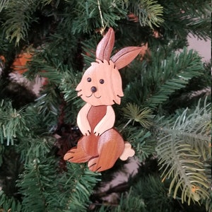 May include: A wooden rabbit ornament with a brown and light brown wood grain finish. The rabbit is sitting with its paws together and has a small tail. The ornament is hanging on a green artificial Christmas tree.