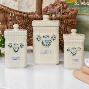 May include: A set of three cream-colored ceramic canisters with lids, each featuring a blue floral design and the French words CAFÉ, FARINE, and THÉ printed in blue. The canisters have a rectangular shape with vertical ribbed detailing.