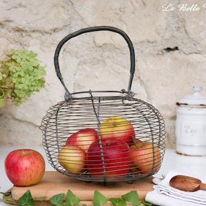 Op de afbeelding: Een draadmand met een handvat bevat verschillende rode en gele appels. De mand staat op een houten snijplank, met een extra appel links. De achtergrond omvat een stenen muur en groen.