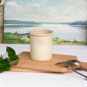 Könnte beinhalten: Ein cremefarbenes Keramikgefäß mit abgerundetem Rand steht auf einem Holzschneidebrett. Im Hintergrund ein Landschaftsbild mit Wasser und Bäumen. Zwei silberne Utensilien liegen auf dem Brett.