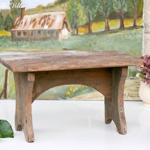 Puede incluir: Un pequeño taburete de madera desgastada con una parte superior rectangular y soportes arqueados. El taburete es de color marrón natural con toques de pintura verde. Una pintura de un paisaje y un jarrón de flores están en el fondo.