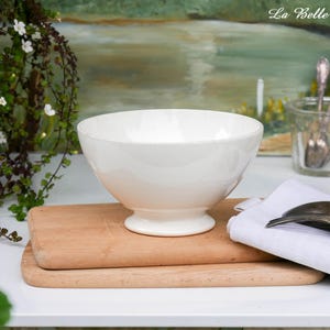 Può includere: Una ciotola in ceramica bianca con base a piedistallo è appoggiata su un tagliere in legno. Un panno bianco e posate sono accanto alla ciotola. Lo sfondo presenta un dipinto e vegetazione.