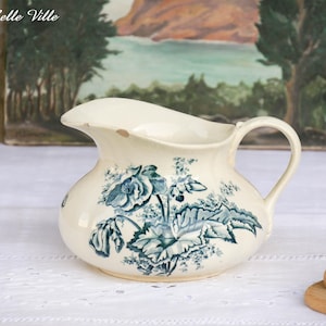 Peut inclure: Pichet en céramique crème avec un motif floral bleu. Le pichet a un bec courbé et une anse. Le motif floral comprend des roses et des feuilles. Le pichet est sur une surface blanche.