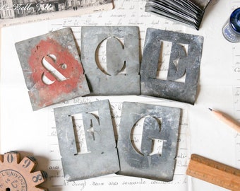 May include: A set of vintage metal letter stencils in various sizes, including the letters C, E, F, and G, and the ampersand symbol. The stencils are painted in shades of gray and red, and have a worn, rustic look.