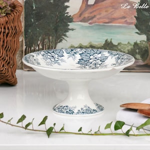 Op de afbeelding: Een witte keramische voetkom met een blauw en wit bloemmotief. De kom heeft een brede, ondiepe schaal en een korte, afgeronde voet. De kom is waarschijnlijk vintage of antiek.