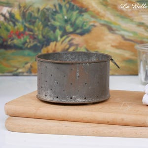 Pode incluir: Um molde cilíndrico de metal com um acabamento cinzento desgastado e uma série de pequenas perfurações espaçadas uniformemente ao longo dos lados. O recipiente possui uma base plana e uma fina aba de metal saliente na borda superior.