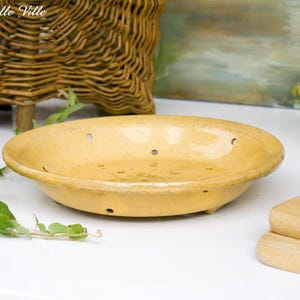 May include: A round, yellow glazed ceramic bowl featuring a series of small, evenly spaced drainage holes along the sides and base. The bowl has a smooth, glossy finish with subtle tonal variations in the yellow glaze and a wide, flat rim.