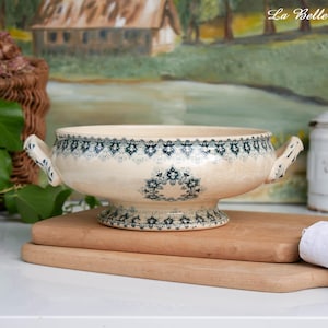 May include: A cream-colored ceramic tureen featuring a dark teal intricate floral pattern around the rim and base. The bowl has two side handles and sits on a pedestal foot. The text La Belle Ville is visible in the upper right corner.
