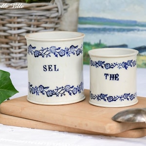 以下が含まれることがあります： 「SEL」と「THE」の文字が入った2つのヴィンテージセラミックキャニスターが木製のボードの上に置かれています。どちらもクリーム色で、青い花の模様とトリムが施されています。銀のスプーンがボードの上に置かれています。