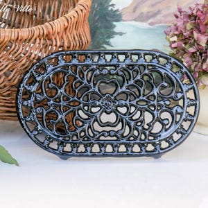 Op de afbeelding: Een ovale, zwarte gietijzeren onderzetter met een sierlijk bloemmotief. De onderzetter is ongeveer 25 cm lang en 15 cm breed. De onderzetter staat op een wit oppervlak, met een rieten mand en een vaas met bloemen op de achtergrond.