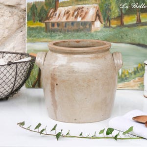 Könnte beinhalten: Ein beigefarbener Steinguttopf mit zwei Griffen, wahrscheinlich für die Küche. Der Topf hat ein rustikales, gealtertes Aussehen mit horizontalen Bändern in der Mitte. Ein Drahtkorb und ein weißes Glas mit Deckel sind ebenfalls zu sehen.