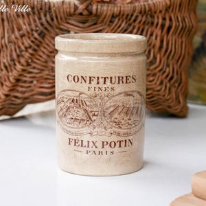 May include: A cylindrical beige ceramic jar featuring brown printed text that reads CONFITURES FINES and FELIX POTIN PARIS, along with two oval illustrations of industrial factory buildings. The jar has a smooth finish and a simple rimmed top.