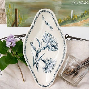 Può includere: Un piatto da portata ovale bianco sporco con un motivo floreale blu e un bordo blu decorativo. Il piatto è posto in un cestino di filo metallico con un rametto di fiori viola e un barattolo di vetro con posate.
