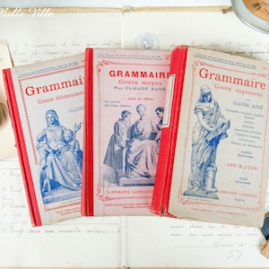 Op de afbeelding: Drie vintage Franse grammatica-leerboeken met rode omslagen en blauwe en witte illustraties. De titels zijn "Grammaire Cours élémentaire", "Grammaire Cours moyen" en "Grammaire Cours supérieur".