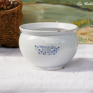 Peut inclure: Un bol en céramique blanche de forme arrondie avec un bord large. L'avant présente une inscription bleue "CONFITURES Nouvelles" entourée d'un petit motif floral décoratif. Le bol a une finition émaillée lisse avec des signes d'usure.