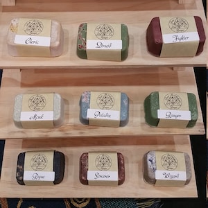 May include: A wooden shelf display with ten bars of soap, each with a different color and design. The soaps are labeled with the names of different Dungeons and Dragons character classes, including Alchemist, Barbarian, Bard, Cleric, Druid, Fighter, Monk, Paladin, Ranger, Rogue, Sorcerer, and Wizard.