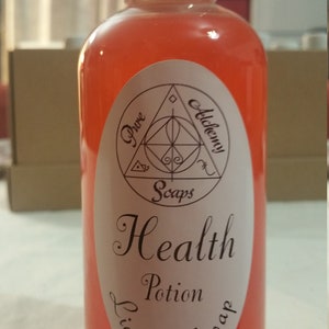 May include: A clear plastic bottle filled with a vibrant orange liquid, topped with a white cap. The bottle features a white label with the words "Health Potion Liquid Soap" and a geometric design. The product is from "Pure Alchemy Soaps."