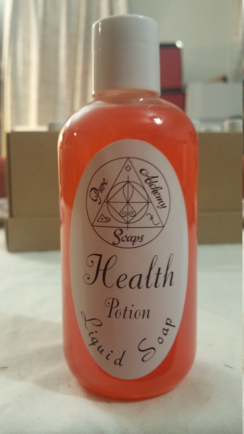 May include: A clear plastic bottle filled with a vibrant orange liquid, topped with a white cap. The bottle features a white label with the words "Health Potion Liquid Soap" and a geometric design. The product is from "Pure Alchemy Soaps."