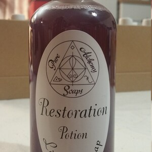 May include: A clear plastic bottle with a white cap, filled with a deep purple liquid soap. The label reads "Pure Alchemy Soaps" and "Restoration Potion Liquid Soap" in elegant script. The bottle is designed for liquid soap.