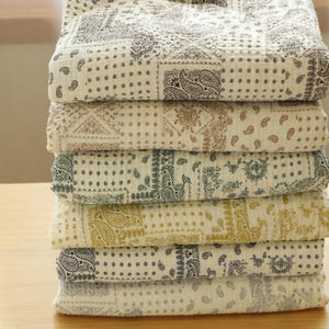 May include: A stack of six folded, patterned blankets. Each blanket features a different color scheme, including cream, tan, and blue, with paisley and polka dot designs. The blankets are stacked on a light brown wooden surface.