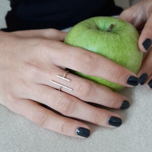 Op de afbeelding: Een zilveren ring met een eenvoudig geometrisch ontwerp. De ring wordt gedragen aan een vinger die een groene appel vasthoudt.