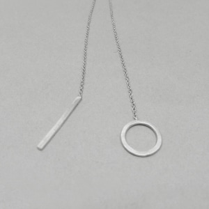 Puede incluir: Un collar de cadena de plata con un colgante de barra largo y delgado y un colgante circular. El collar está sobre un fondo blanco.
