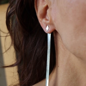 Könnte beinhalten: Silberne Ohrringe mit einem diamantförmigen Ohrstecker oben und einer langen, dünnen Silberkette, die herunterhängt.