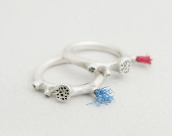 Puede incluir: Dos anillos de plata con un diseño texturizado y un pequeño punto negro en el centro. Los anillos están conectados por una cuerda azul y roja.