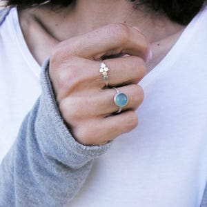 Puede incluir: Dos anillos de plata en una mano. El anillo en el dedo índice tiene una piedra preciosa azul claro. El anillo en el dedo medio tiene una fila de pequeños círculos de plata.